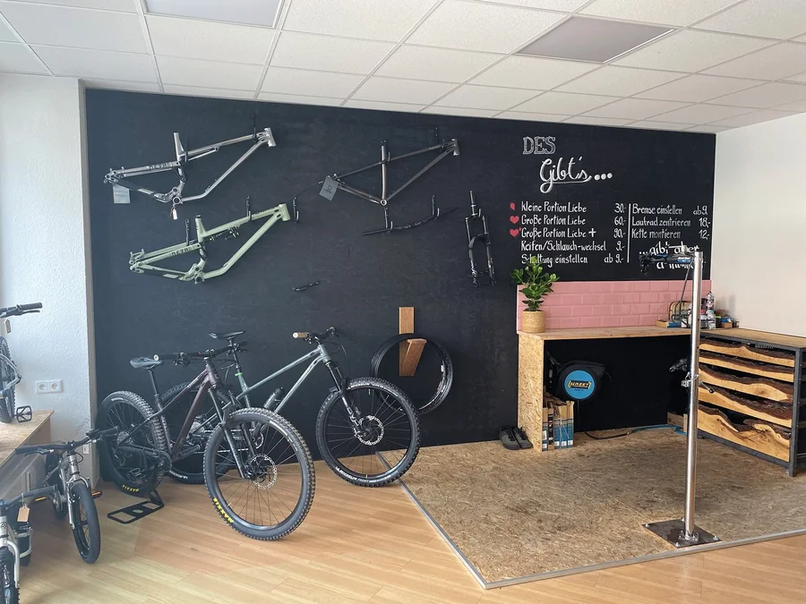 Die Werkstatt im Fahrradschuppen: Rahmen an der schwarzen Wand, Räder, Montageständer und die Tafel mit den Preisen